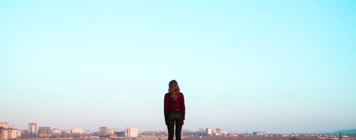 A woman stands alone in a vast open field, gazing towards a distant city skyline under a clear blue sky.
