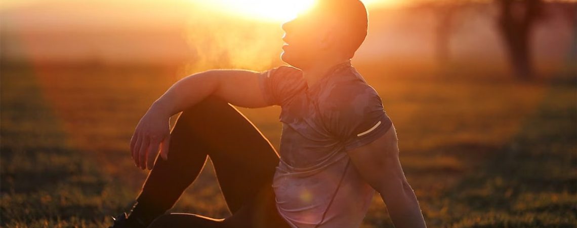 Muscular man sits in rural field at sunset, exhaling visible breath.